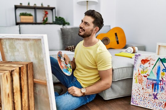 Young man with beard painting canvas at home looking away to side with smile on face, natural expression. laughing confident.