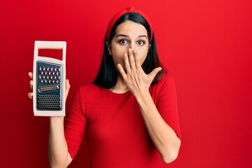 Young hispanic woman holding grater covering mouth with hand, shocked and afraid for mistake....