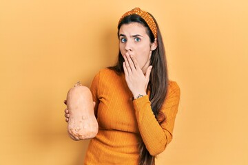 Young brunette teenager holding healthy fresh pumpkin covering mouth with hand, shocked and afraid...