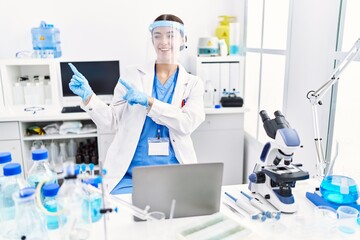 Young hispanic woman wearing scientist uniform smiling and looking at the camera pointing with two hands and fingers to the side.