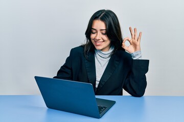 Beautiful hispanic woman working at the office with laptop doing ok sign with fingers, smiling friendly gesturing excellent symbol