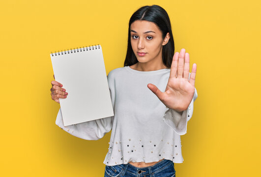 Young hispanic girl holding notebook with open hand doing stop sign with serious and confident expression, defense gesture