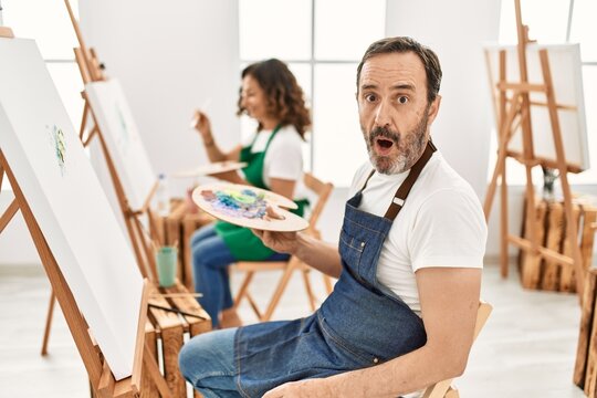 Hispanic middle age man and mature woman at art studio scared and amazed with open mouth for surprise, disbelief face