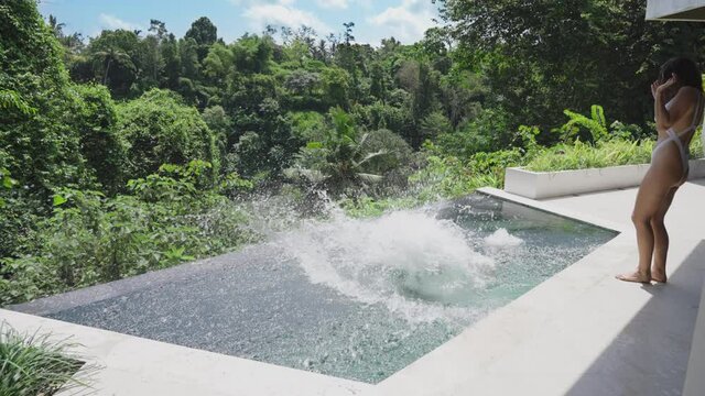 A young man in a bathrobe stands at the side of the pool and touches the water with his foot, a girl approaches him and pushes him into the pool. Guy grabs girl by the leg and pulls her into the pool 