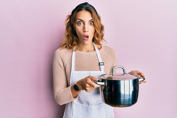 Young caucasian woman wearing apron holding cooking pot afraid and shocked with surprise and amazed...