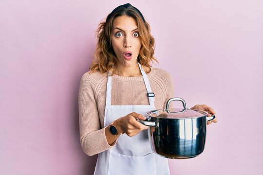 Young caucasian woman wearing apron holding cooking pot afraid and shocked with surprise and amazed expression, fear and excited face.