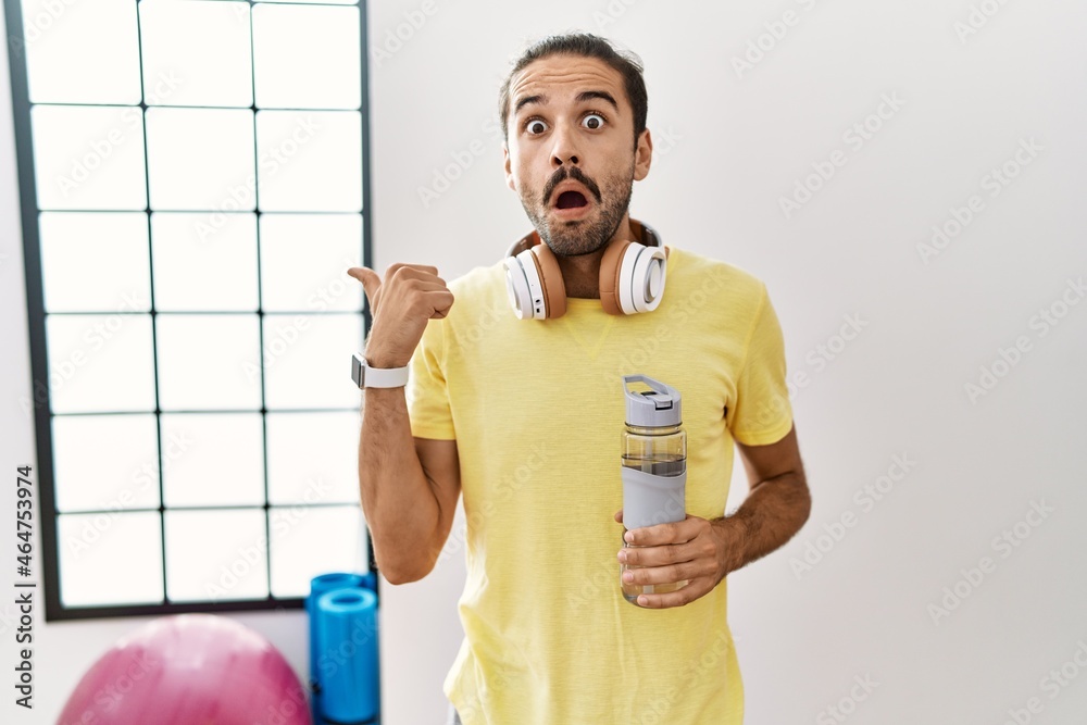 Sticker young hispanic man wearing sportswear and drinking water at the gym surprised pointing with hand fin