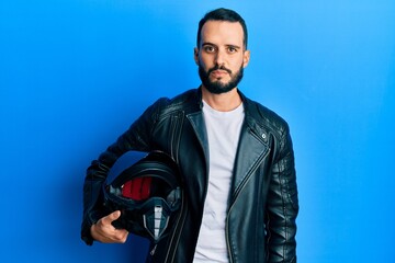 Young man with beard holding motorcycle helmet thinking attitude and sober expression looking self confident