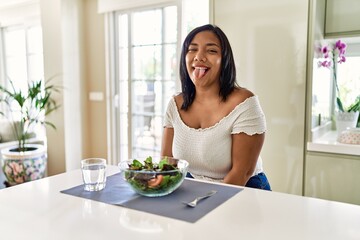 Obraz na płótnie Canvas Young hispanic woman eating healthy salad at home sticking tongue out happy with funny expression. emotion concept.
