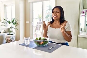 Obraz na płótnie Canvas Young hispanic woman eating healthy salad at home gesturing finger crossed smiling with hope and eyes closed. luck and superstitious concept.