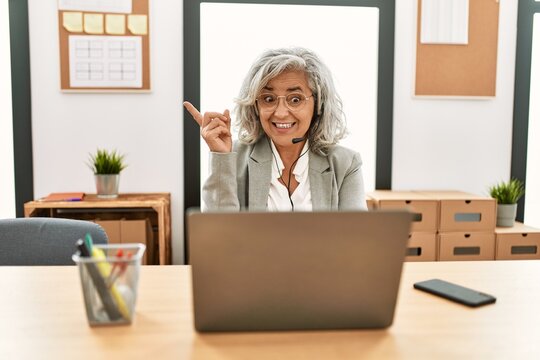 Middle age businesswoman sitting on desk working using laptop at office with a big smile on face, pointing with hand and finger to the side looking at the camera.