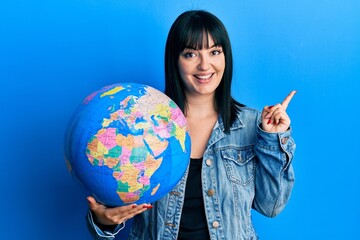 Young hispanic woman holding world ball smiling happy pointing with hand and finger to the side