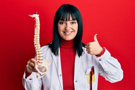 Young hispanic doctor woman holding anatomical model of spinal column smiling happy and positive, thumb up doing excellent and approval sign