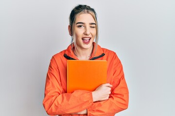 Young modern girl holding book winking looking at the camera with sexy expression, cheerful and...