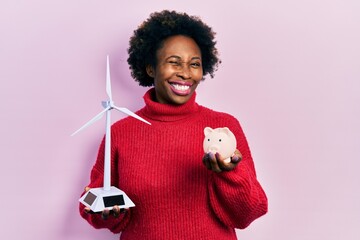 Young african american woman holding windmill and piggy bank winking looking at the camera with...