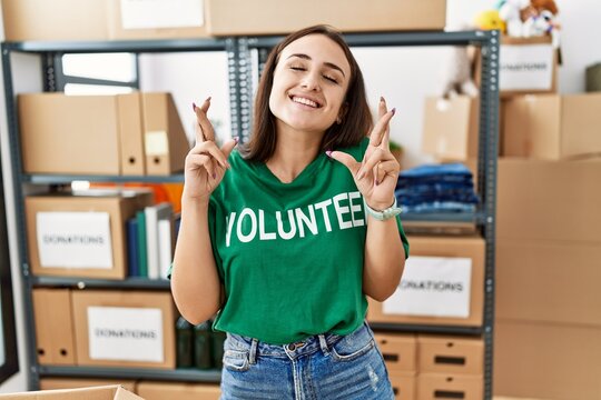 Young brunette woman wearing volunteer t shirt at donations stand gesturing finger crossed smiling with hope and eyes closed. luck and superstitious concept.