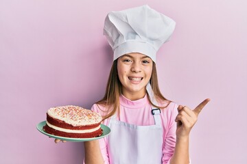 Beautiful brunette little girl wearing baker uniform holding homemade cake smiling happy pointing with hand and finger to the side