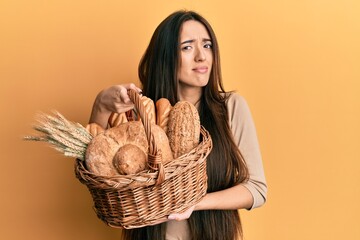 Young hispanic girl holding wicker basket with bread clueless and confused expression. doubt...