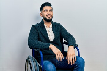 Handsome man with beard sitting on wheelchair looking away to side with smile on face, natural...