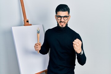 Handsome man with beard holding brushes close to easel stand angry and mad raising fist frustrated and furious while shouting with anger. rage and aggressive concept.