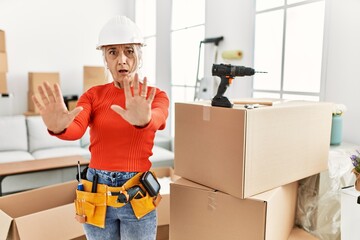 Middle age grey-haired woman wearing hardhat standing at new home afraid and terrified with fear expression stop gesture with hands, shouting in shock. panic concept.