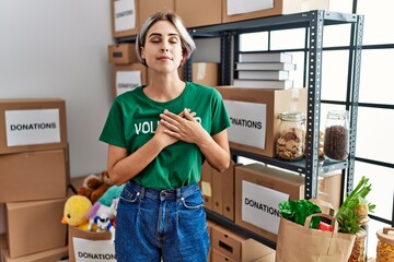 Young beautiful woman wearing volunteer t shirt at donations stand smiling with hands on chest with...