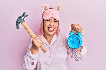 Hispanic woman with pink hair wearing pajama holding hammer and alarm clock winking looking at the...