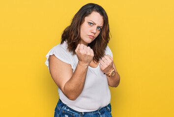 Young plus size woman wearing casual white t shirt ready to fight with fist defense gesture, angry and upset face, afraid of problem