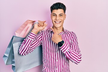 Young hispanic man holding shopping bags smiling happy pointing with hand and finger