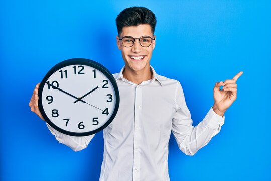 Young hispanic man holding big clock smiling happy pointing with hand and finger to the side