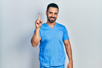 Naklejka premium Handsome hispanic man with beard wearing blue male nurse uniform showing and pointing up with finger number one while smiling confident and happy.