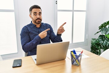 Young hispanic man with beard working at the office with laptop pointing aside worried and nervous with both hands, concerned and surprised expression