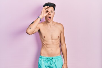 Fototapeta premium Young hispanic man wearing swimwear shirtless doing ok gesture shocked with surprised face, eye looking through fingers. unbelieving expression.