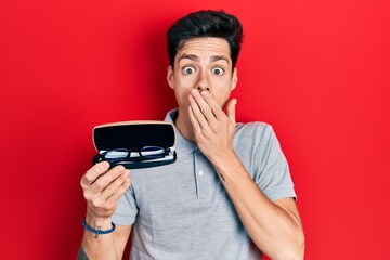 Young hispanic man holding glasses covering mouth with hand, shocked and afraid for mistake....