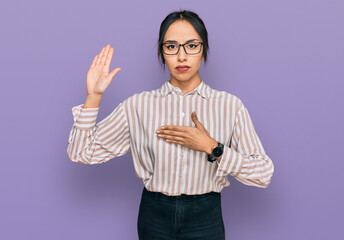 Young hispanic girl wearing casual clothes and glasses swearing with hand on chest and open palm,...