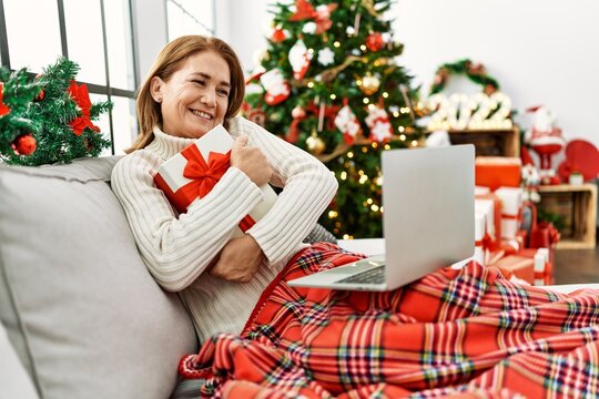 Middle age caucasian woman using laptop holding gift sitting by christmas tree at home