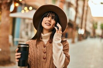 Brunette woman wearing winter hat speaking on the phone and drinking a cup of coffee outdoors at the city