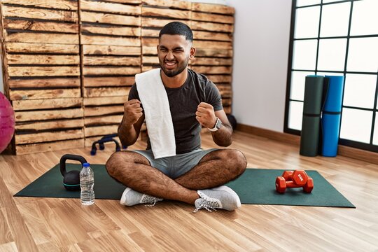 Young indian man sitting on training mat at the gym very happy and excited doing winner gesture with arms raised, smiling and screaming for success. celebration concept.