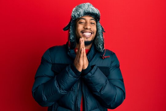Young african american man with beard wearing winter hat and coat praying with hands together asking for forgiveness smiling confident.