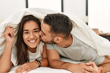 Fototapeta premium Young latin couple kissing and covering with bed sheet lying on the bed.