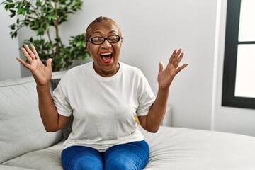 Mature hispanic woman sitting on the sofa at home celebrating mad and crazy for success with arms...