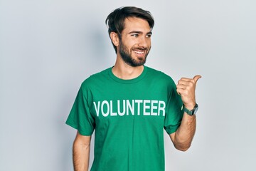 Young hispanic man wearing volunteer t shirt smiling with happy face looking and pointing to the...