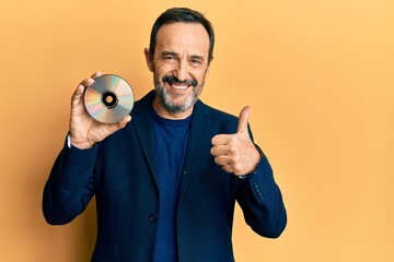 Middle age hispanic man holding compact disc smiling happy and positive, thumb up doing excellent...