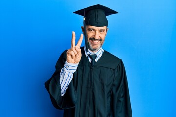 Middle age hispanic man wearing graduation cap and ceremony robe showing and pointing up with fingers number two while smiling confident and happy.