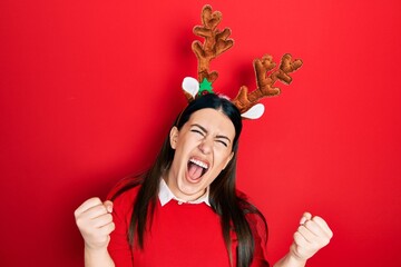 Young hispanic woman wearing cute christmas reindeer horns screaming proud, celebrating victory and...