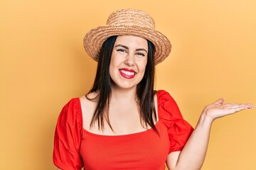 Young hispanic woman wearing summer hat smiling cheerful presenting and pointing with palm of hand looking at the camera.