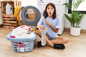 Young hispanic girl doing laundry holding socks surprised pointing with finger to the side, open...