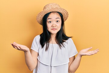 Young chinese girl wearing summer hat clueless and confused expression with arms and hands raised....