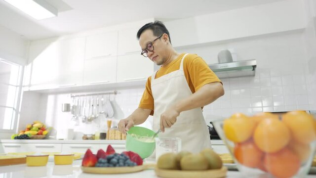Asian man bakery shop owner preparing bakery in the kitchen. Adult male making whipping cream from fresh milk in the bowl. Small business entrepreneur and indoor activity lifestyle baking concept.