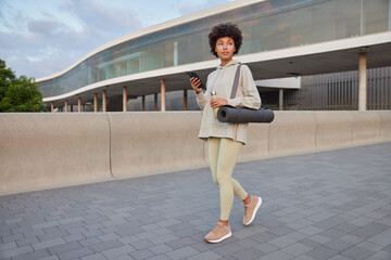 Sporty motivated woman in activewear carries karemat holds bottle with water and smartphone checks receieved notifications whilehaving workout walks outdoors has regular training. Healthy lifestyle